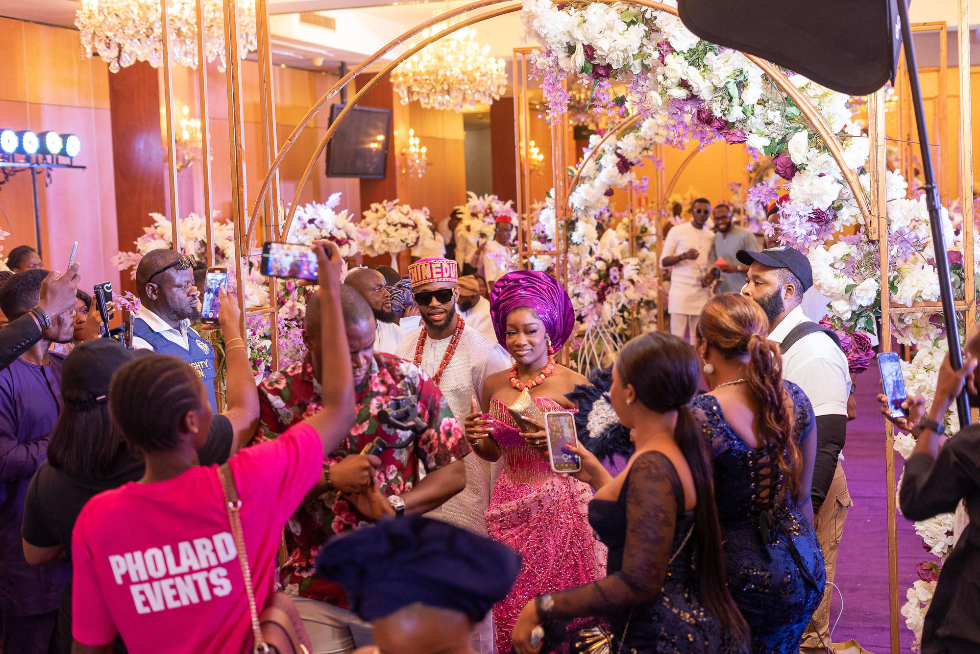 Couple dressed in traditional attire walking through a floral archway while guests photograph and celebrate them at an indoor event.