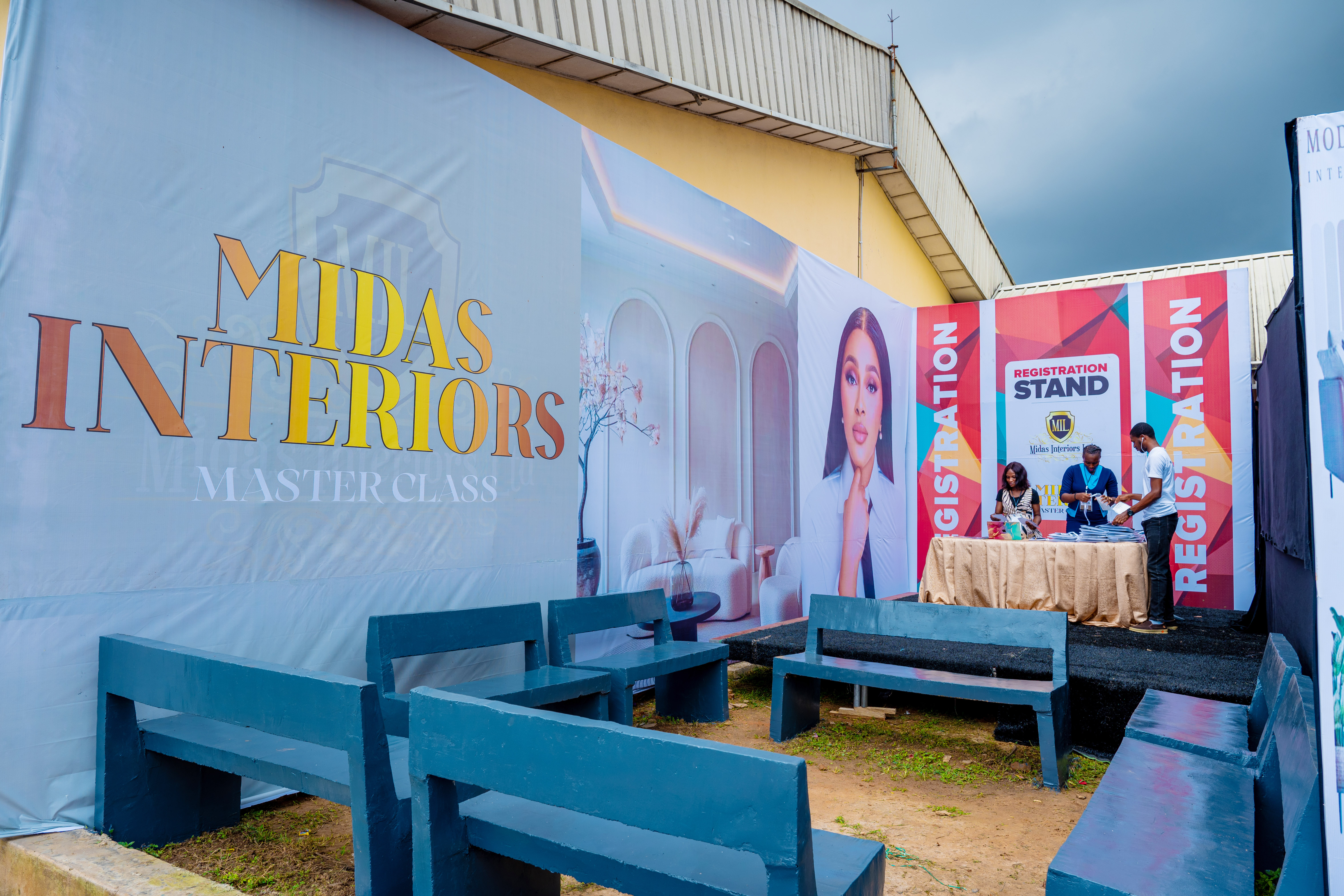 Outdoor registration stand for Midas Interiors Master Class with three people at a table and blue benches arranged for seating.