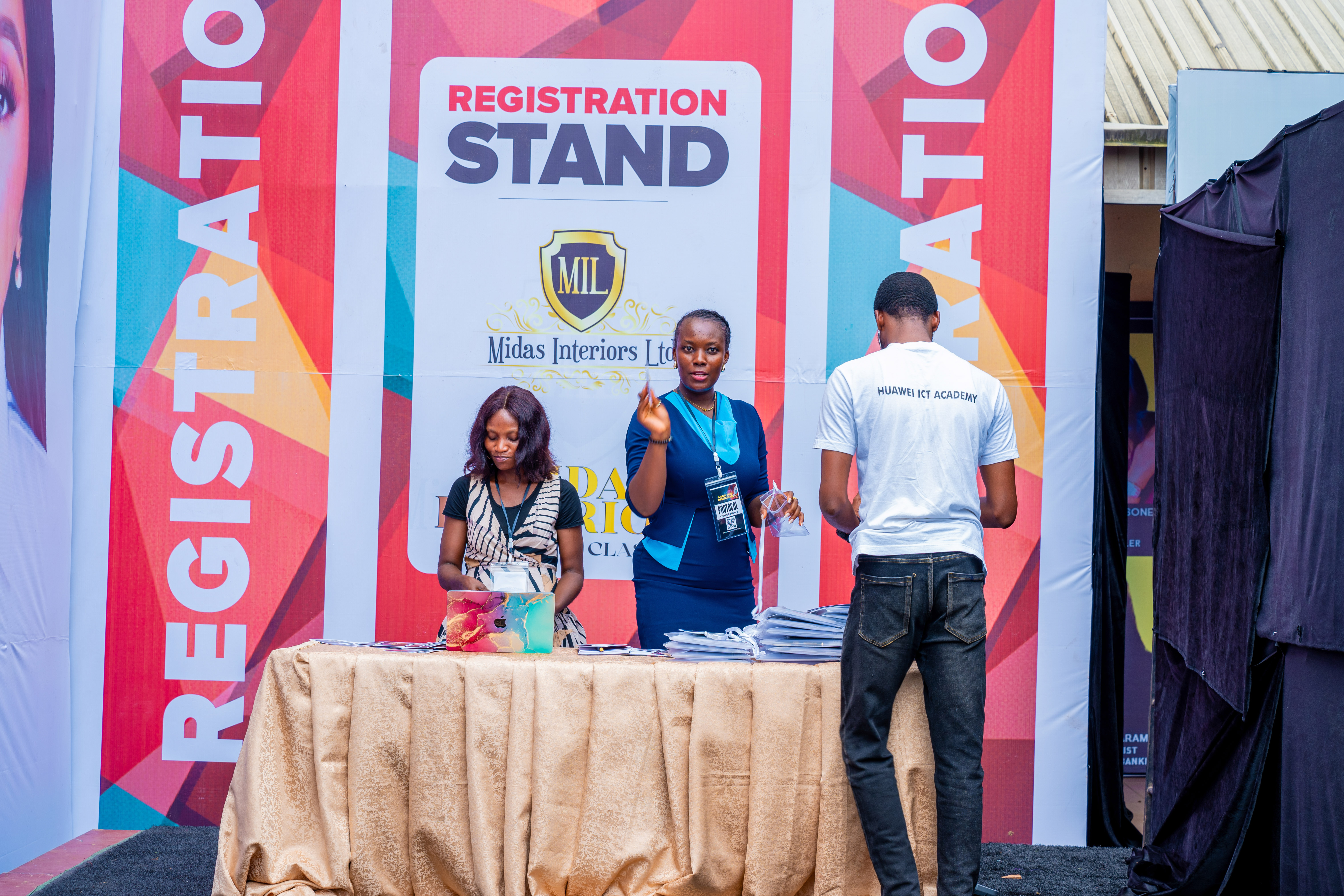 Three people at a registration stand with two women behind a table and a man in a Huawei ICT Academy shirt standing in front.