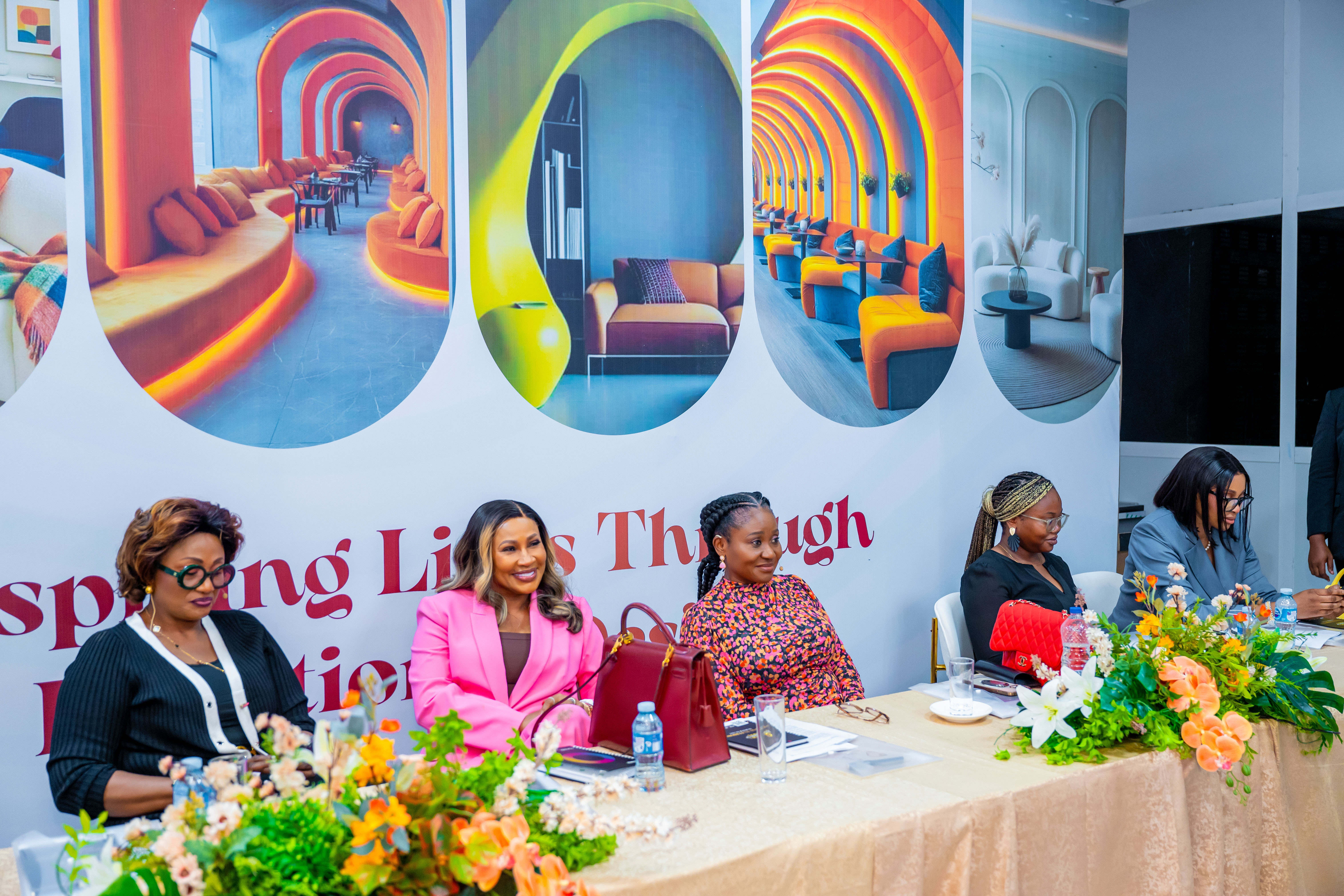 Five women seated at a table with floral arrangements in front and colorful modern interior photos displayed behind them.