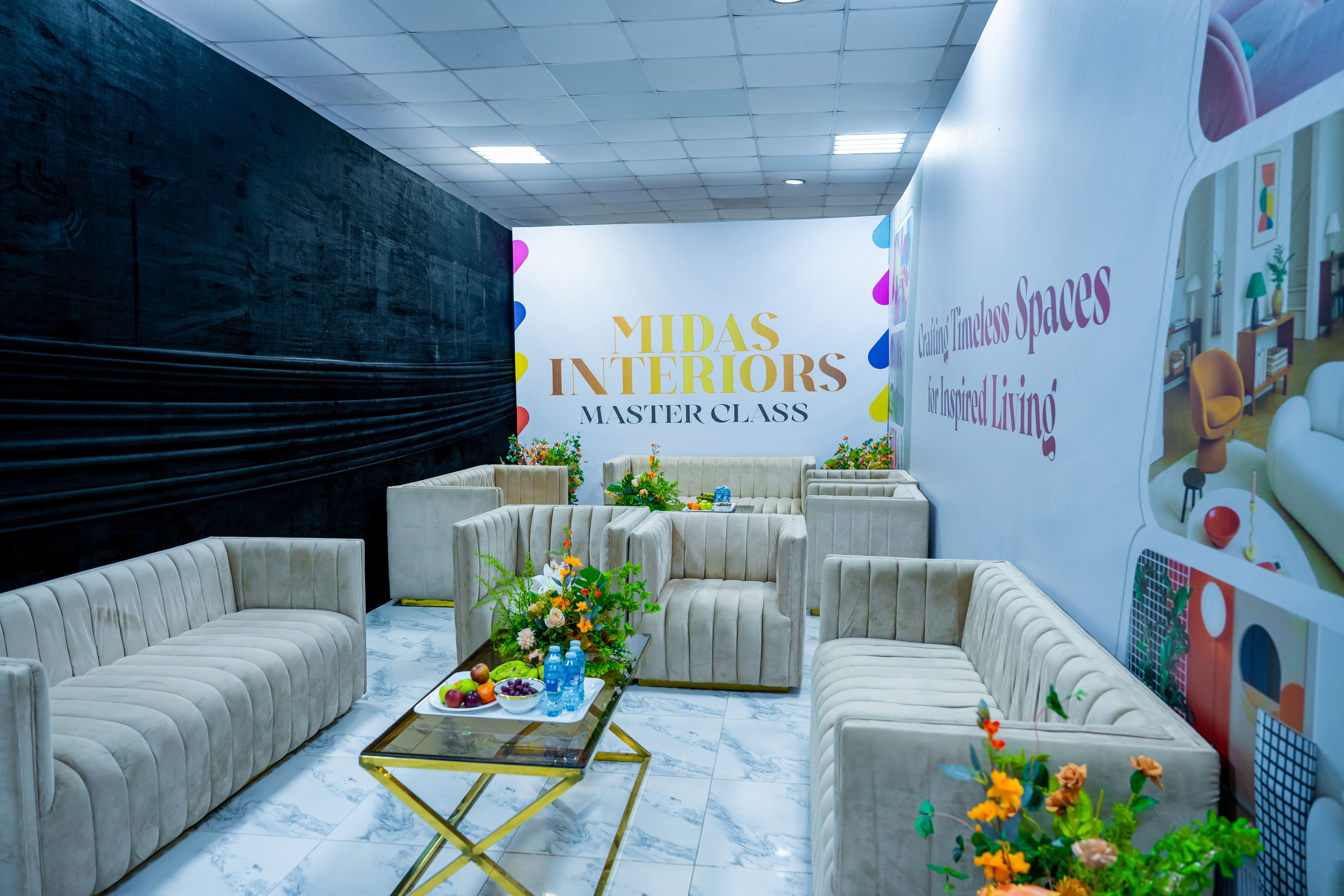 Modern interior room with beige velvet sofas arranged around a glass coffee table with fruit, water bottles, and floral arrangements, featuring a sign on the back wall that reads 'MIDAS INTERIORS MASTER CLASS.'