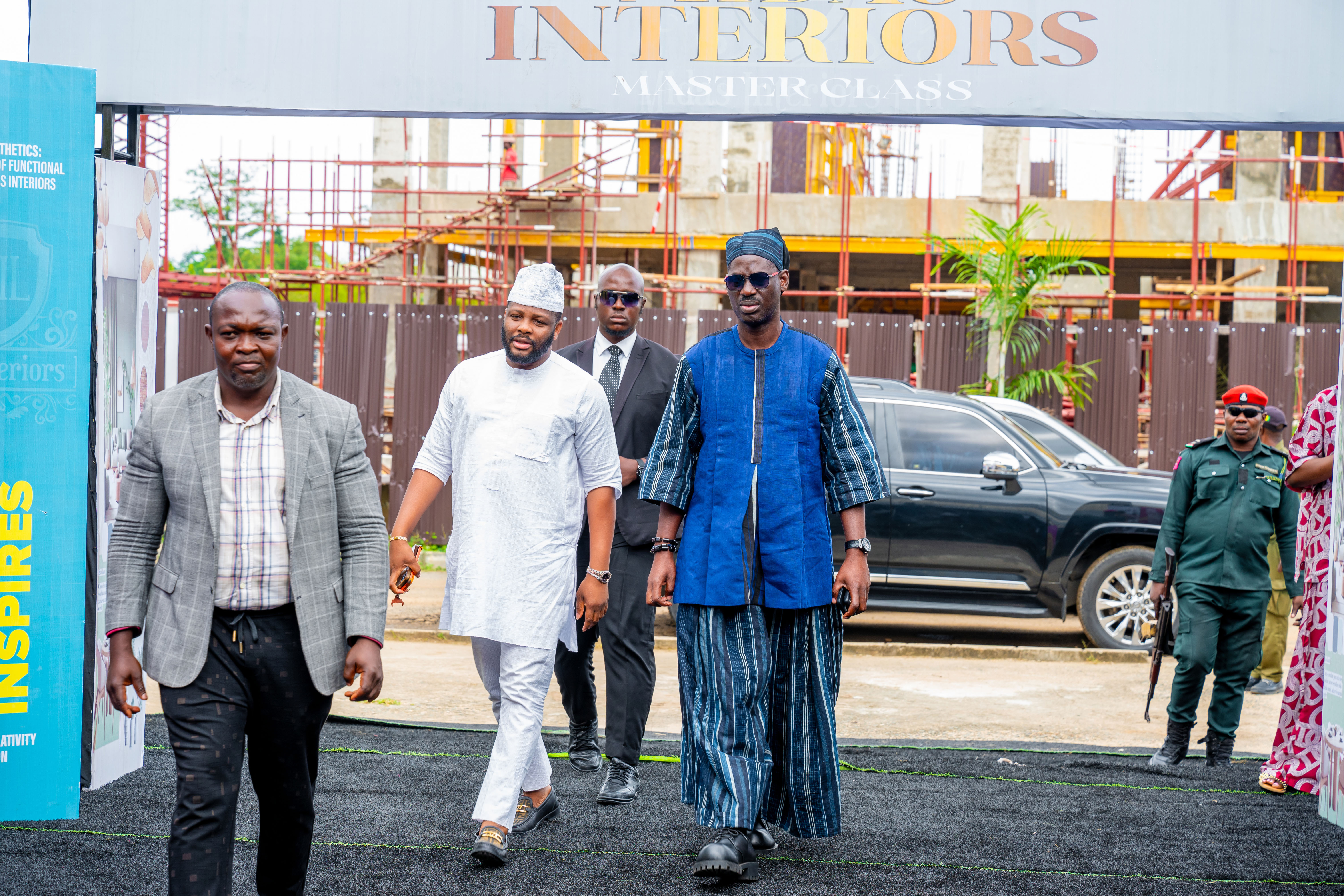 Four men walking on a black carpet outdoors with a construction site and a black SUV in the background; one man in traditional blue attire, another in white, one in a gray blazer, and a security officer in uniform.