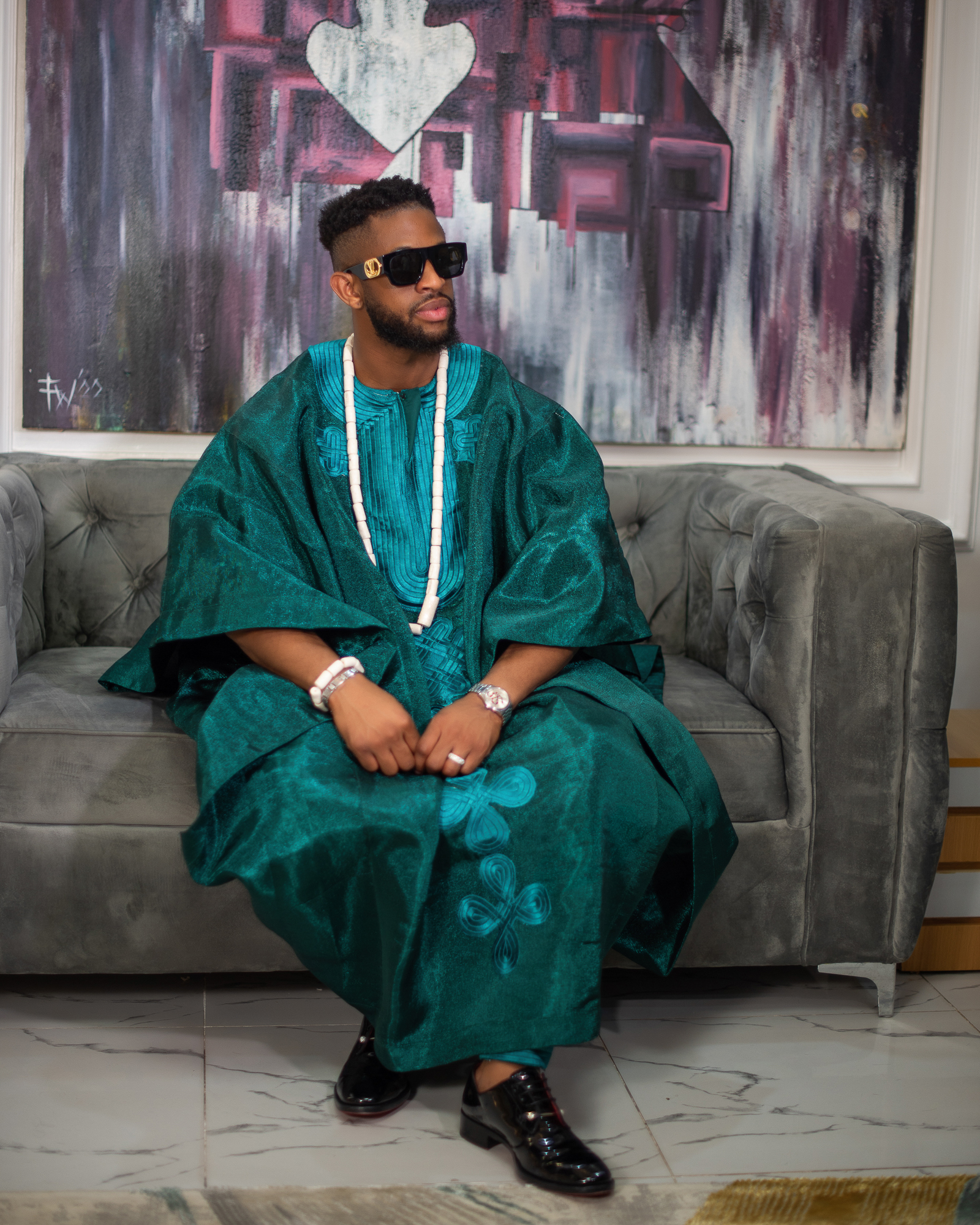 Man wearing traditional teal agbada attire, sunglasses, and beaded necklace sitting on a gray tufted sofa.