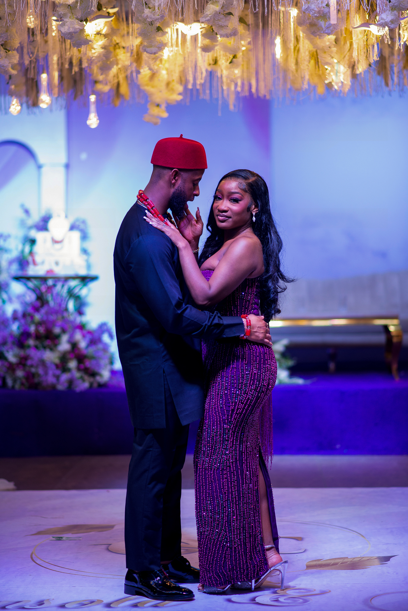 Couple dressed elegantly, embracing and posing indoors under hanging floral decorations and warm lights.