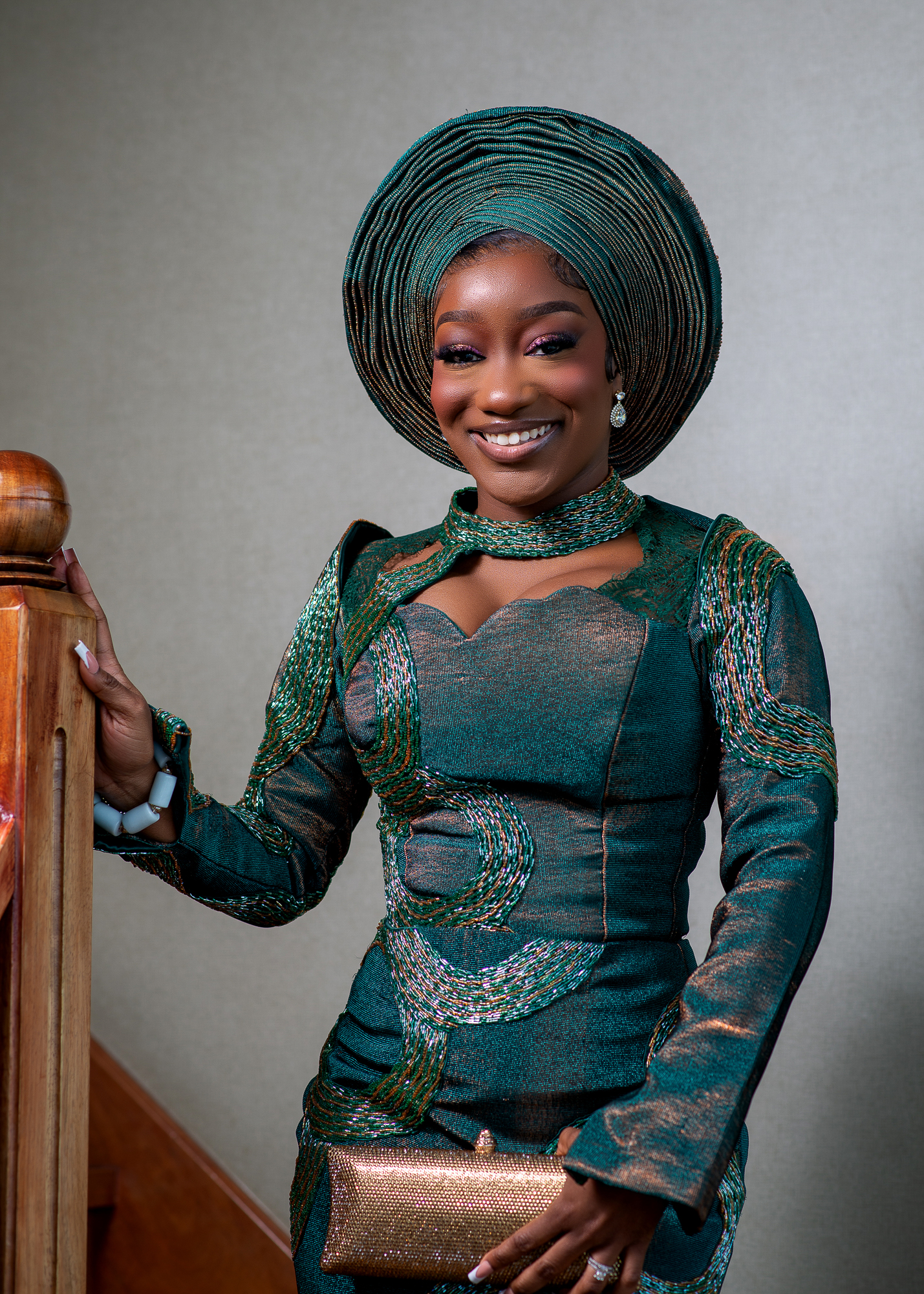 Smiling woman in traditional green and gold attire with matching headwrap, holding a gold clutch and standing by a wooden railing.
