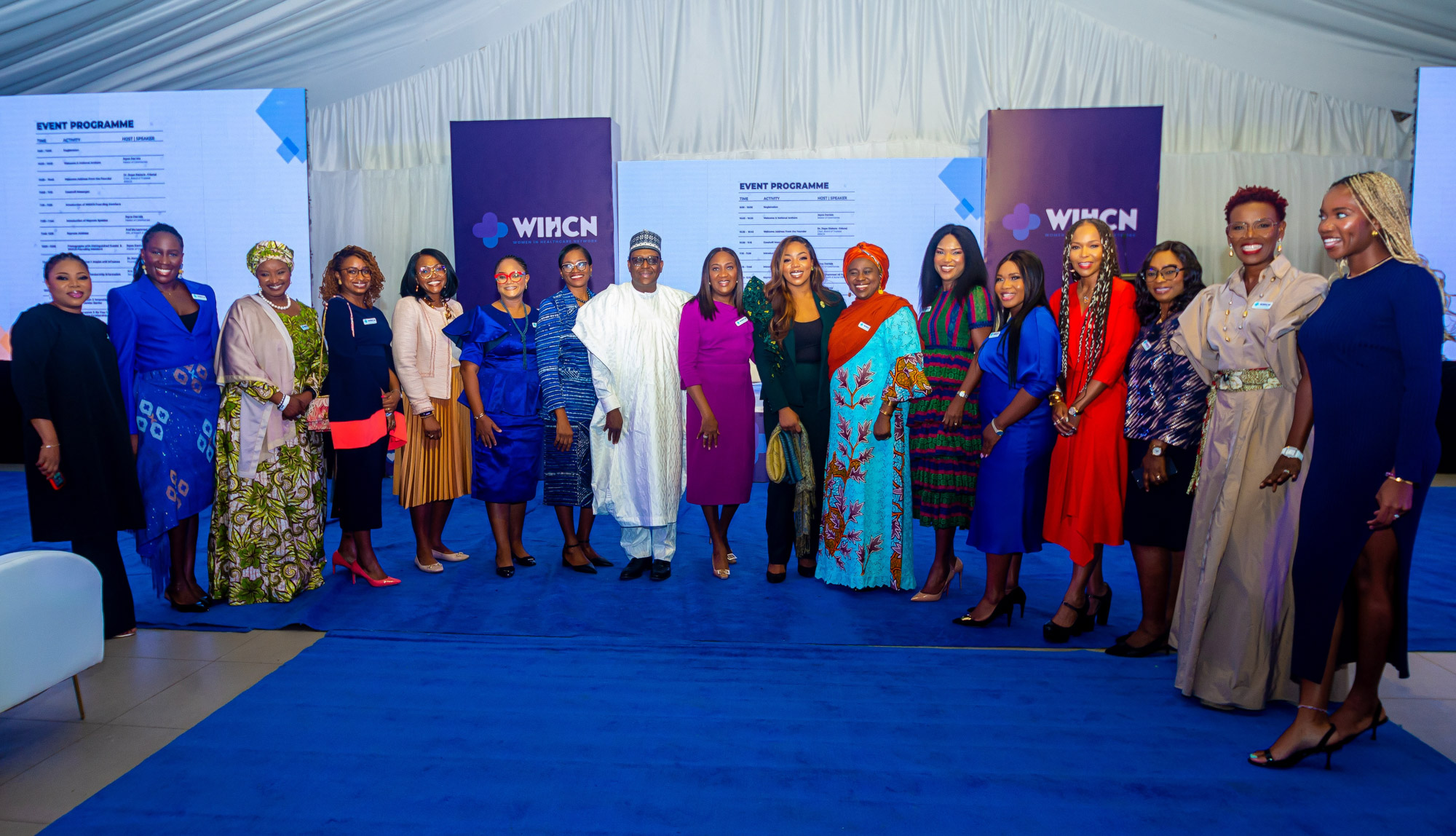 Group of sixteen people, including one man in traditional white attire, standing in a row on a blue carpet at a WIHCN event with event programme screens behind them.