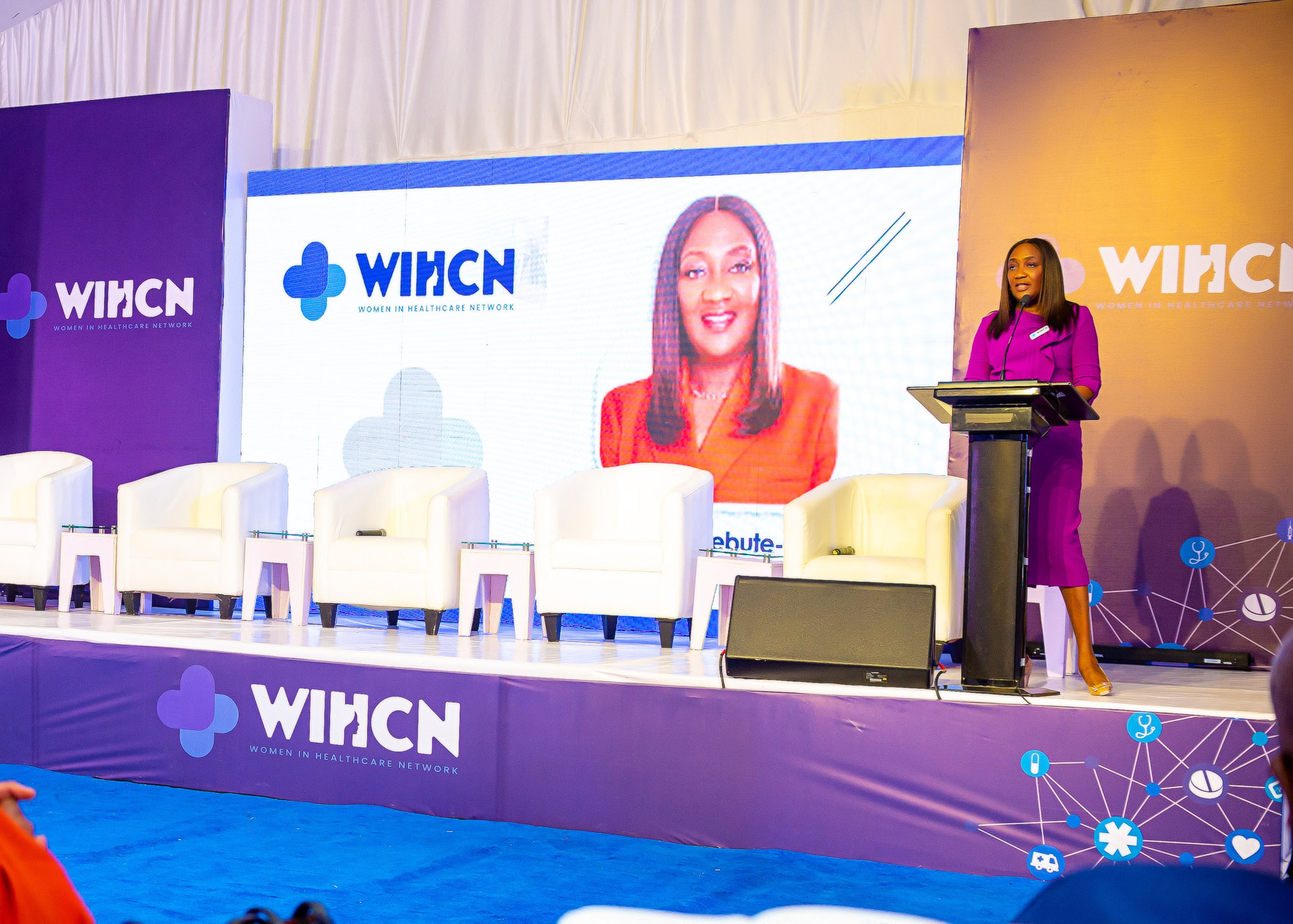 Woman in purple dress speaking at a podium on a stage with white chairs and Women in Healthcare Network banners.