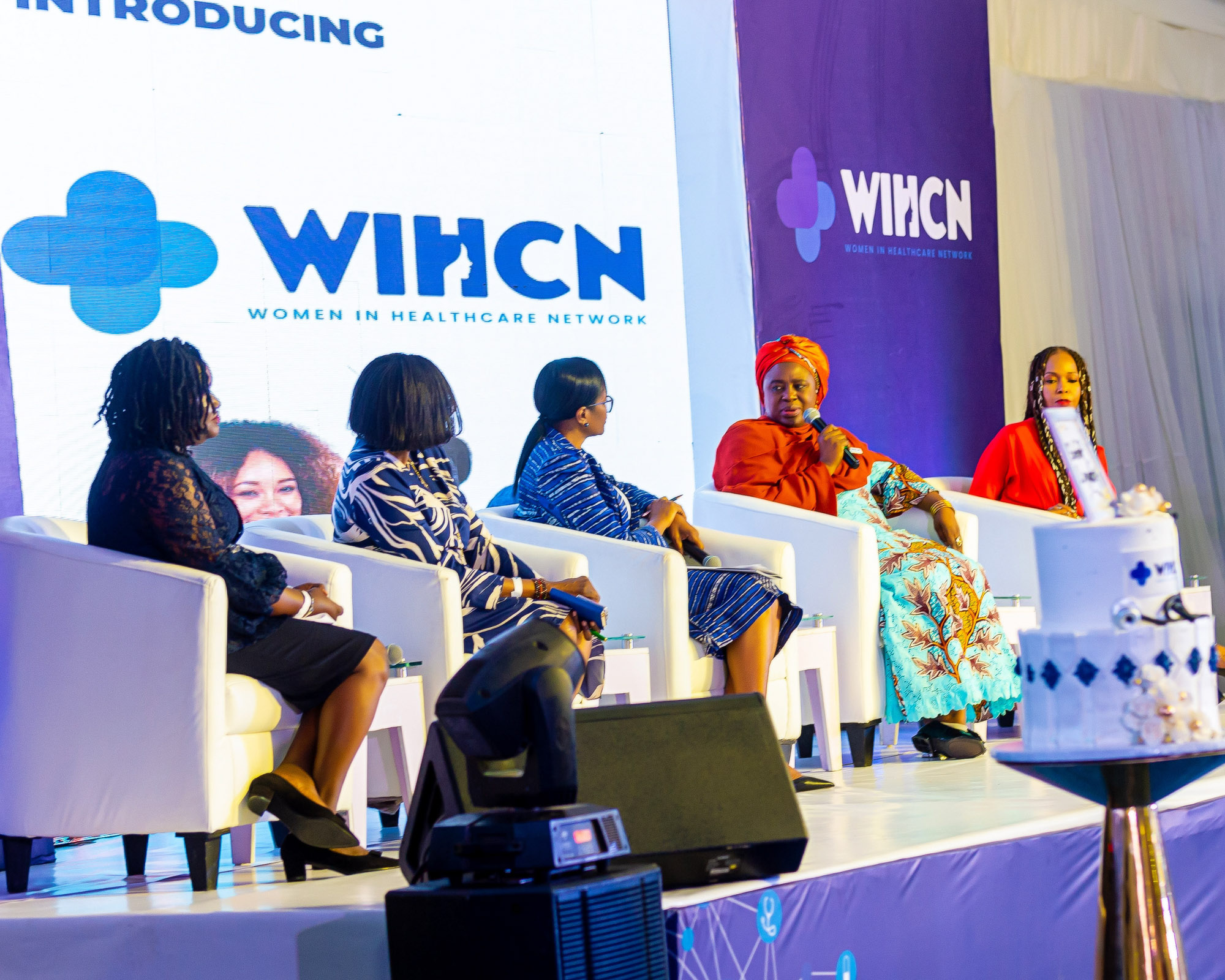 Panel of five women seated on a stage during a Women in Healthcare Network event, with a branded backdrop and a decorated cake in the foreground.