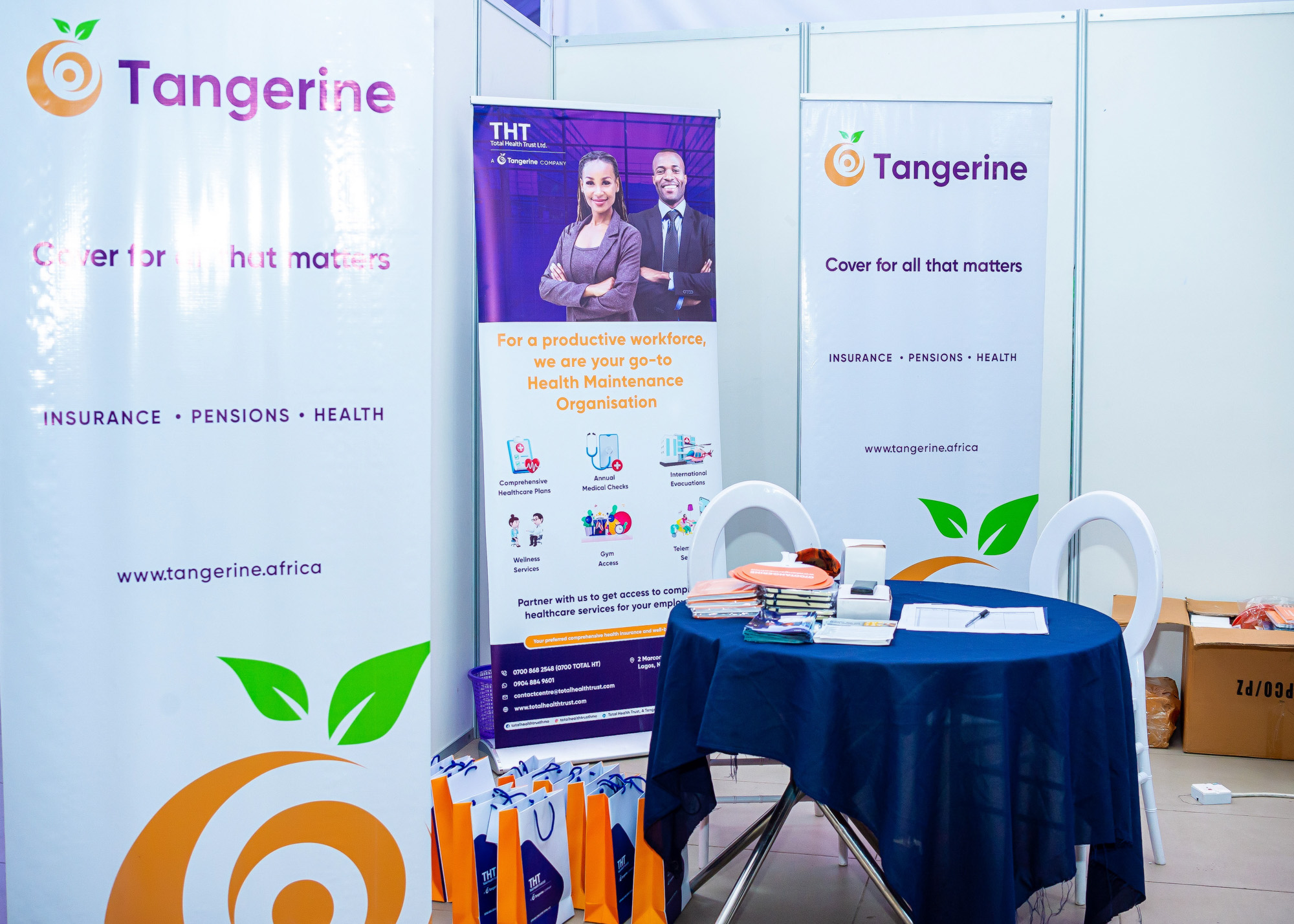 Booth setup with Tangerine and THT banners, a round table covered with a dark blue cloth, two white chairs, brochures, and gift bags on the floor.