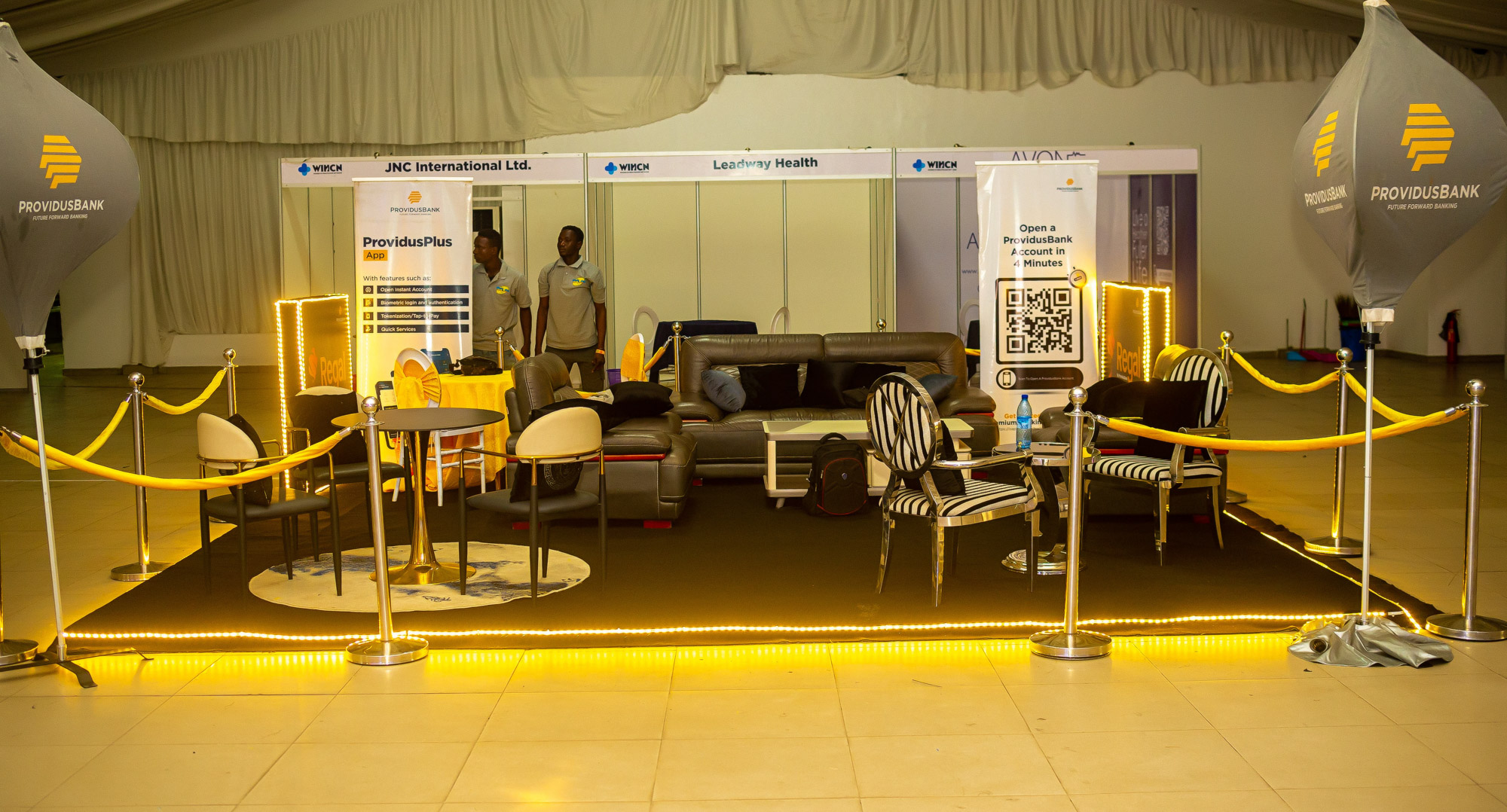 ProvidusBank promotional booth with sofas, striped chairs, informational banners, and yellow rope barriers illuminated by yellow lights.