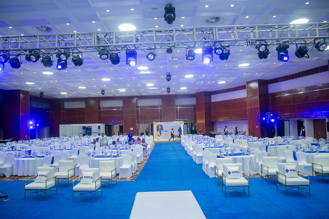 Large conference hall with rows of round tables covered in white cloths, white chairs, and a blue carpet aisle under bright ceiling lights and stage lighting.