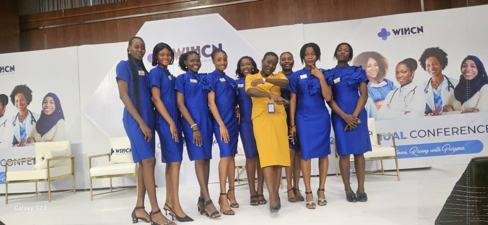 Group of nine women standing on a stage, eight wearing matching blue dresses and one in a yellow dress, posing and smiling at an event with WHCN branding in the background.