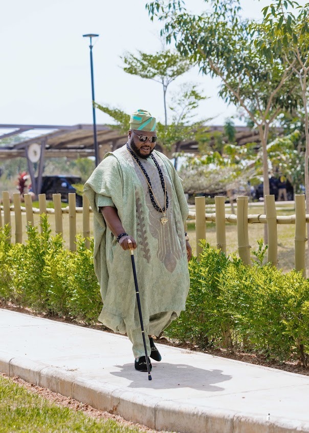 Man dressed in traditional green agbada attire with matching cap and black walking cane, walking on a paved path beside greenery.