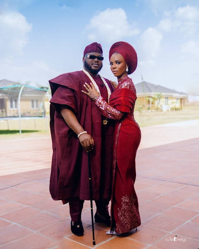Couple dressed in matching traditional Nigerian attire, standing outdoors on a tiled patio under a partly cloudy sky.