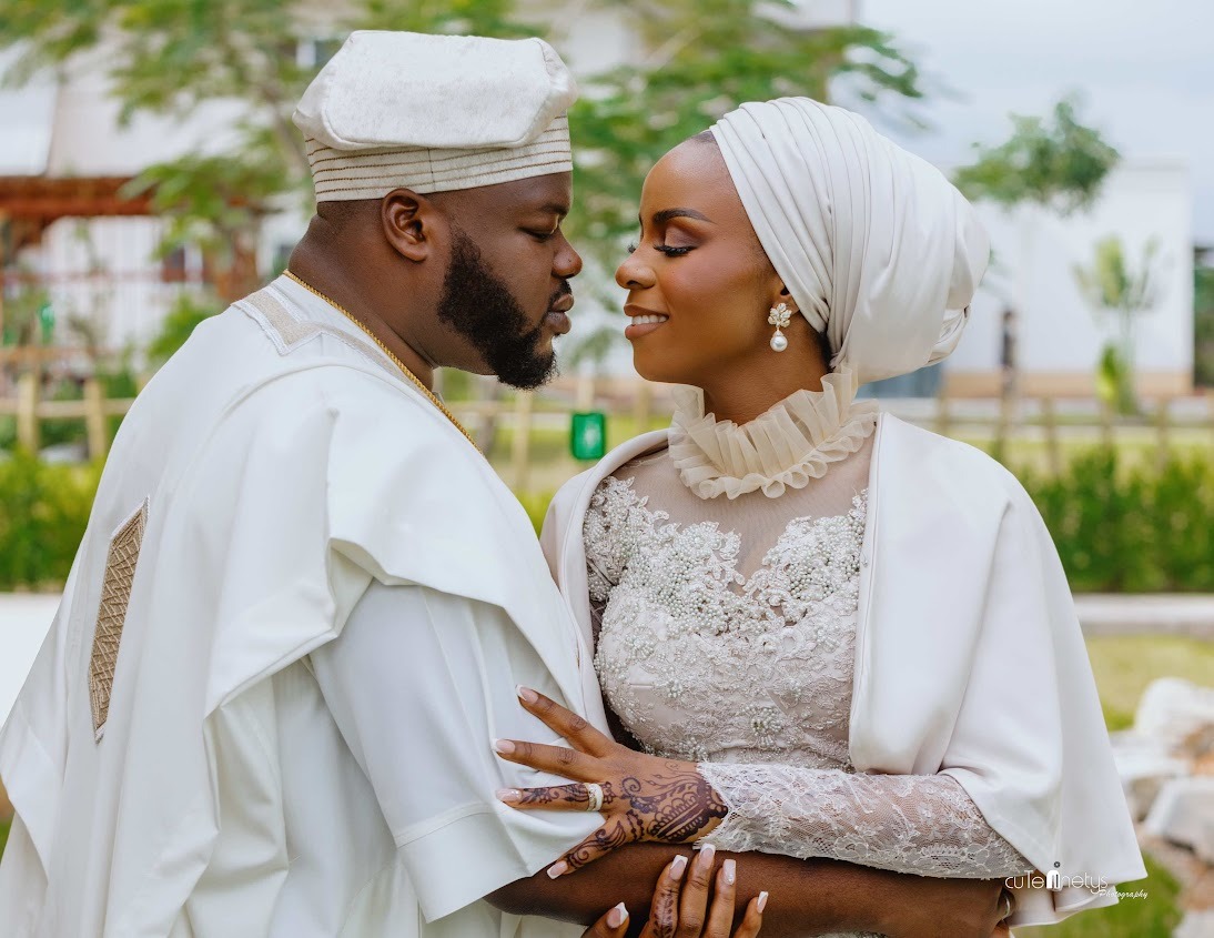 Couple dressed in traditional white attire embracing closely outdoors, with the woman wearing a detailed lace dress and headwrap, and the man wearing a matching cap.