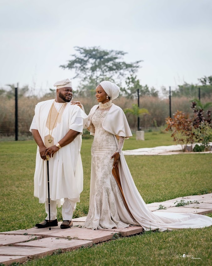 Couple dressed in traditional white attire standing on a stone path in a grassy garden; the woman wears a long gown with a headwrap, and the man holds a cane.