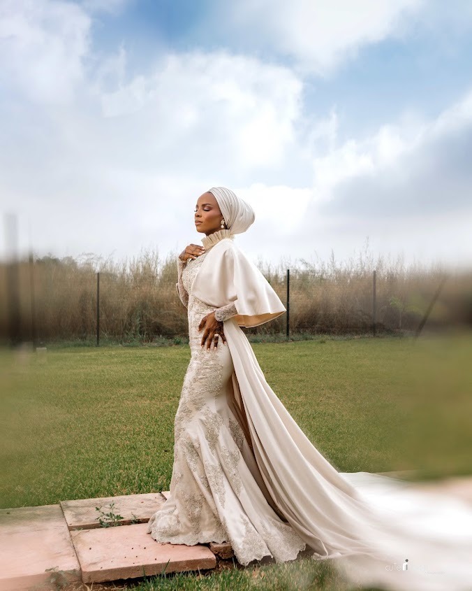 Woman in elegant white lace dress and matching headwrap standing on stone tiles with grassy field and cloudy sky background.