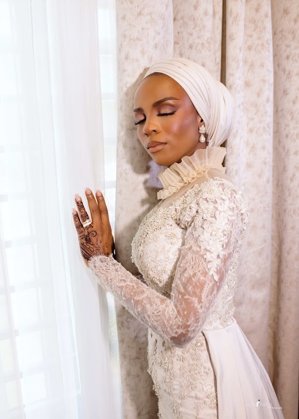 Woman in elegant white lace dress and matching headscarf with henna on hand, standing peacefully with eyes closed near a window.