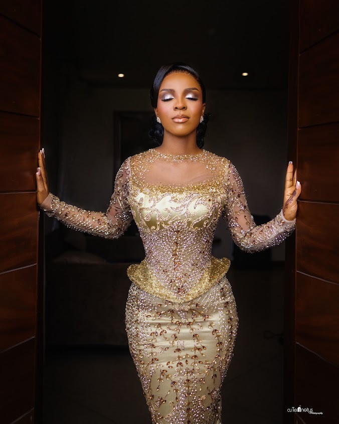 Woman standing in a doorway with closed eyes, wearing an elegant gold dress embellished with intricate beadwork.