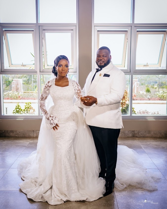 Bride in a lace long-sleeve white wedding gown and groom in a white tuxedo jacket and black pants standing hand in hand indoors in front of large windows.