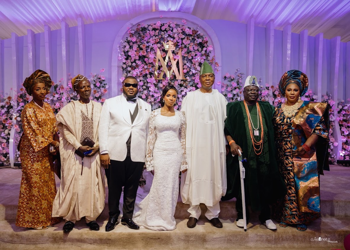 A bride and groom standing with four adults in traditional Nigerian attire in front of a pink floral backdrop at a wedding.