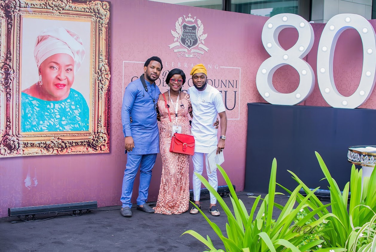 Three people posing at an 80th birthday celebration event with a large framed portrait of a woman and illuminated number 80 in the background.