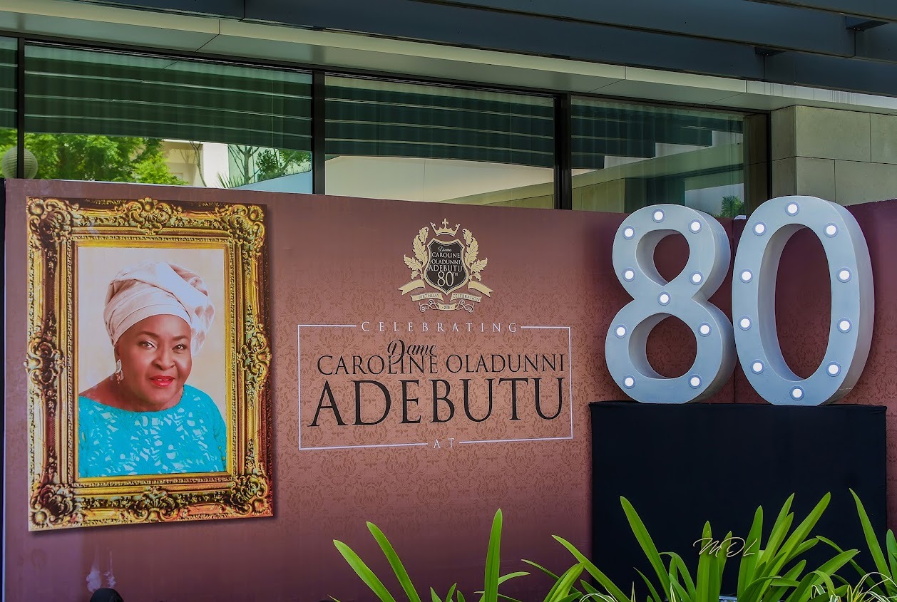 Banner celebrating Dame Caroline Oladunni Adebuto's 80th birthday with her portrait and large lighted number 80.