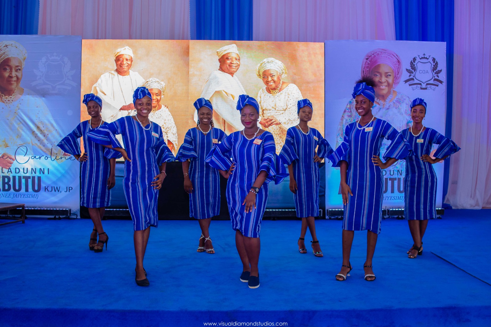Seven women in blue striped dresses and matching headwraps posing on a blue stage with large portraits as a backdrop.