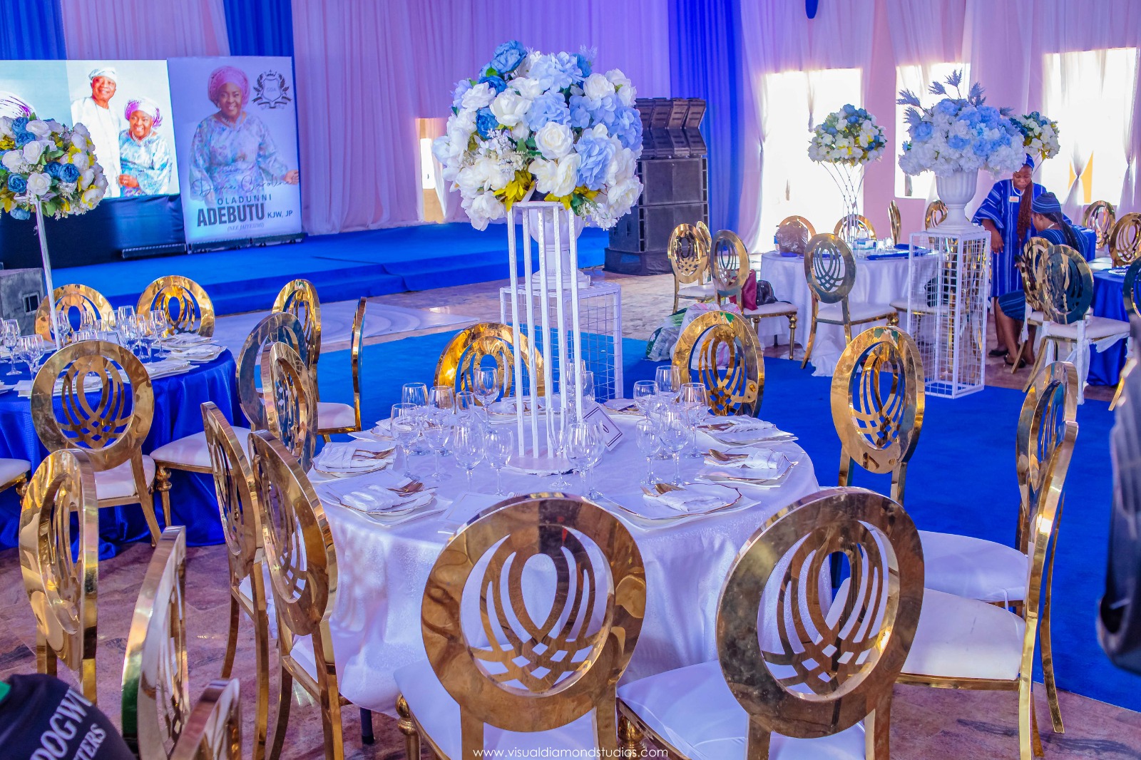 Elegant event setup with round tables covered in white and blue linens, gold decorative chairs, and tall floral centerpieces of white and blue flowers.
