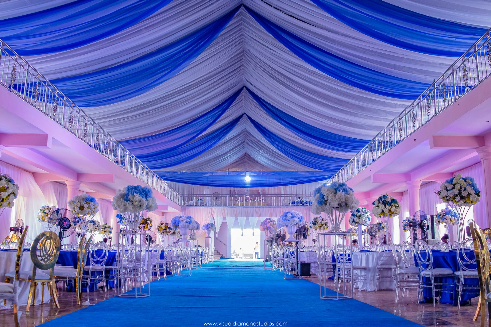 Elegant event hall decorated with blue and white draped ceiling, lined tables with floral centerpieces, and transparent chairs.