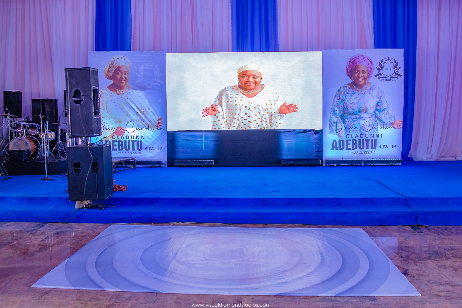Event stage with blue carpet and large screens displaying a woman in traditional attire, flanked by banners with her photo and name Dame Caroline Oladunni Adebutu.