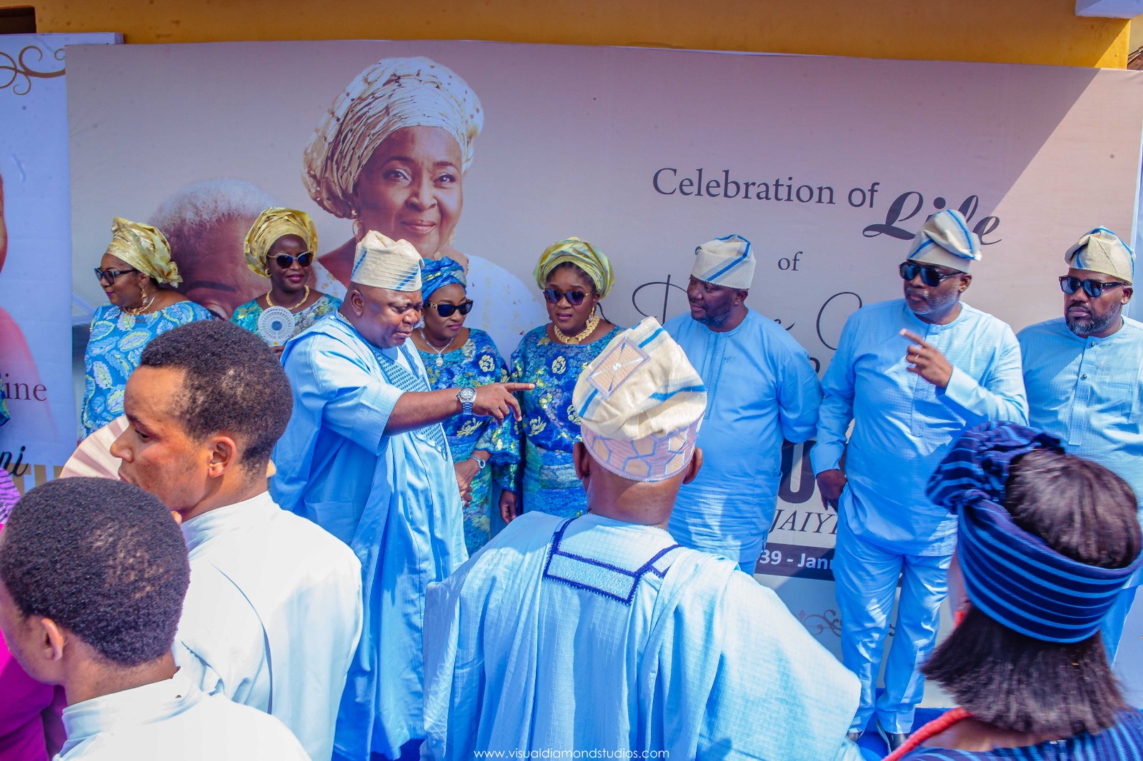 Group of people dressed in traditional Nigerian attire in front of a large banner reading 'Celebration of Life'.