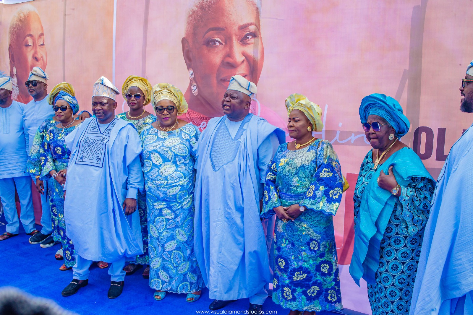 Group of people dressed in vibrant traditional blue and yellow Nigerian attire posing together indoors against a pink backdrop with a large portrait.