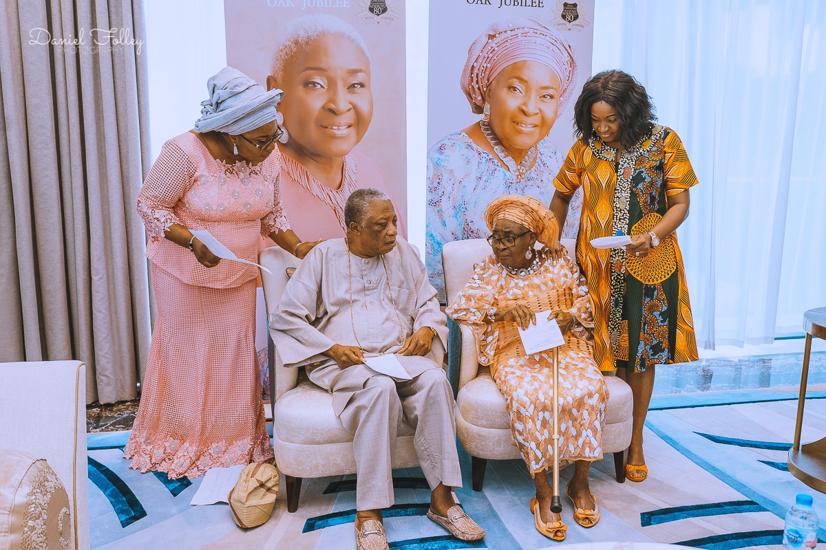 Two elderly individuals seated in chairs holding papers, flanked by two women standing beside them, set against banners featuring portraits and text reading Oak Jubilee.