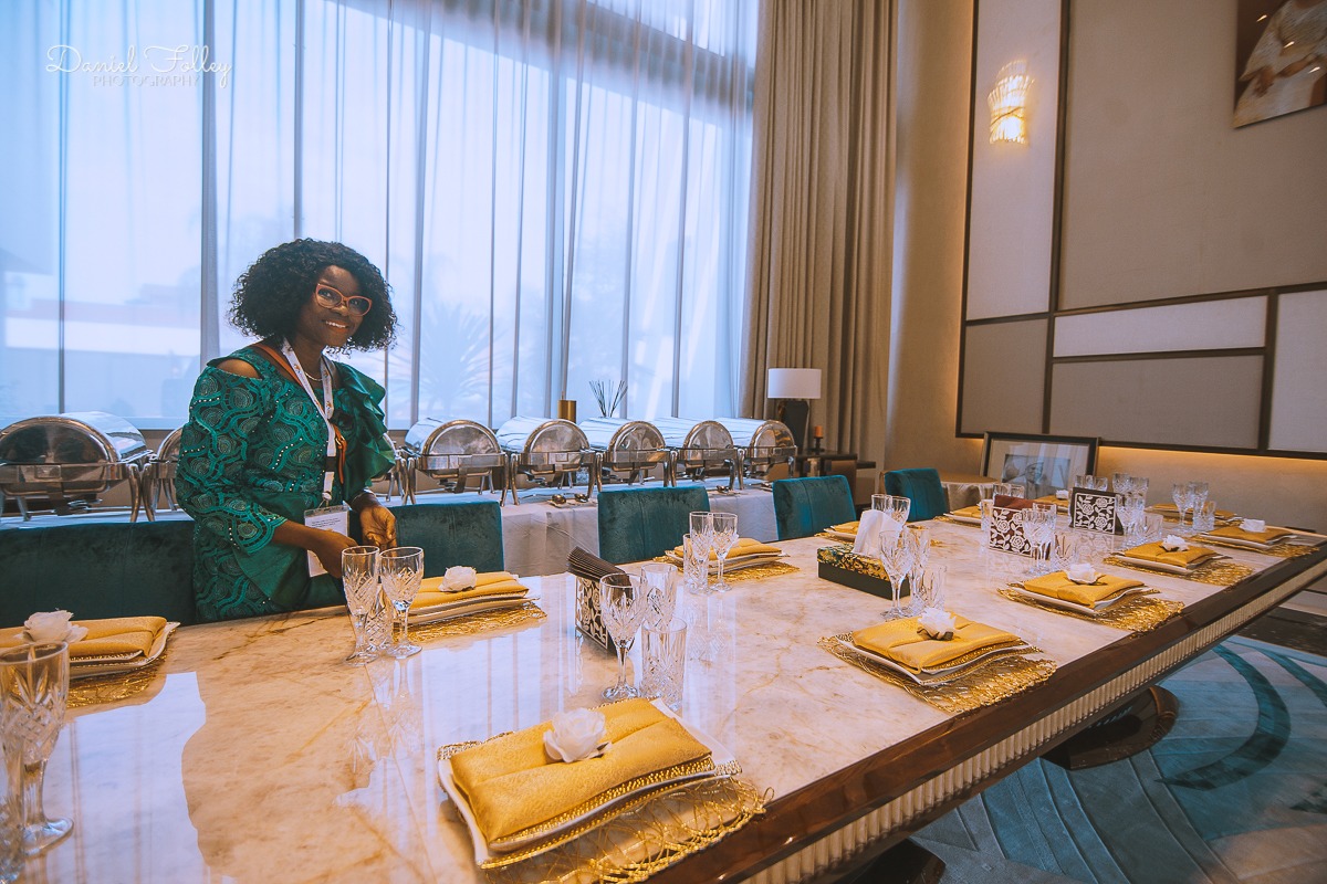 Woman in a green dress standing by a long, elegant dining table set with yellow napkins, crystal glasses, and gold placemats in a well-lit banquet room.