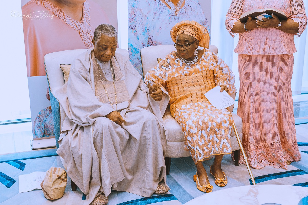Elderly man and woman seated together, holding hands; woman wears a patterned orange dress and headwrap and holds a cane, man in traditional attire.