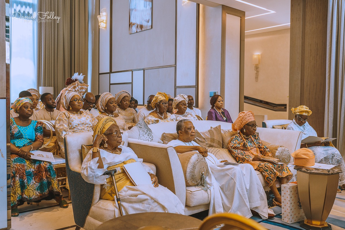 Group of elderly and middle-aged people seated indoors, dressed in colorful traditional African attire, attentively listening.