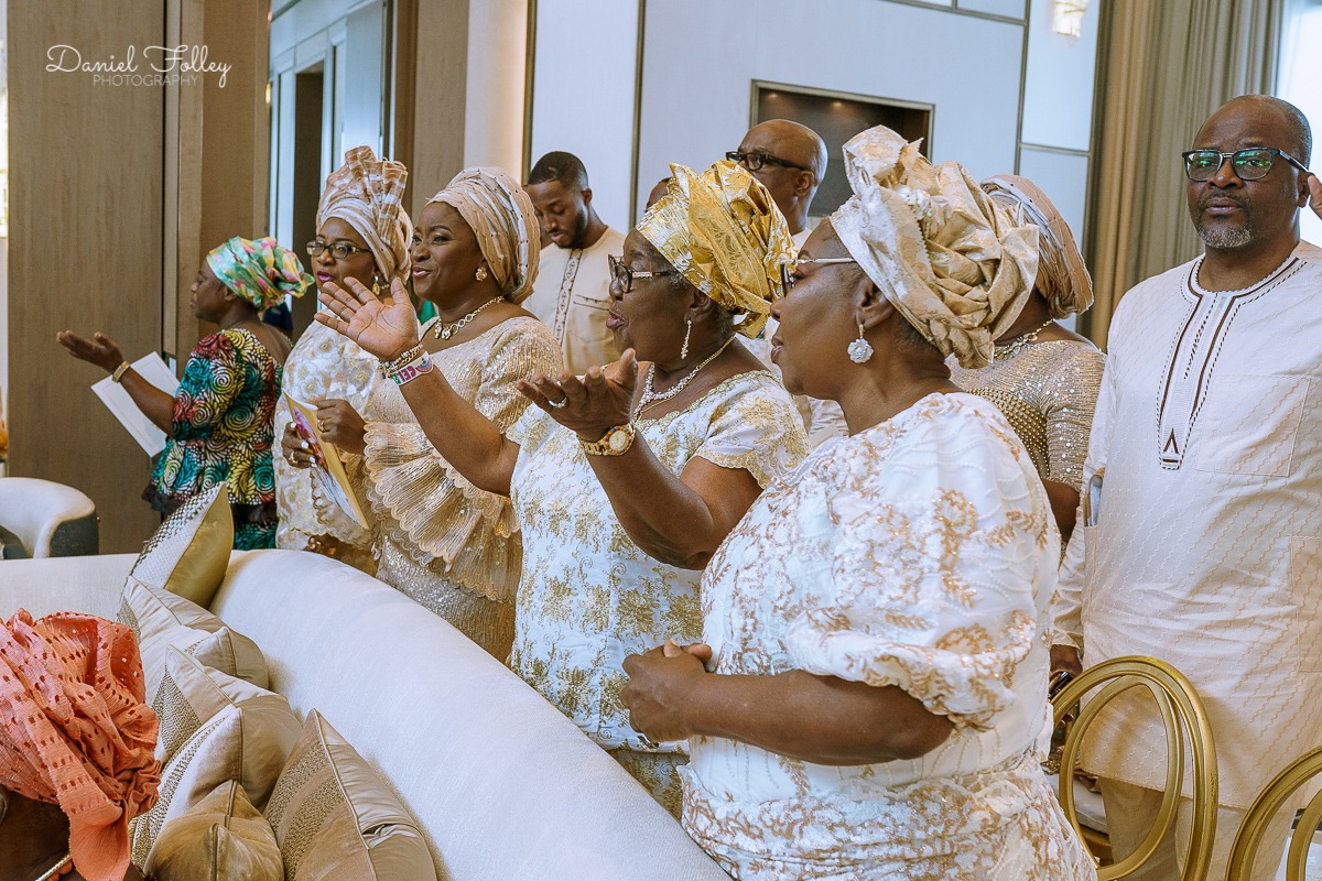 Group of people dressed in traditional African attire standing indoors, some with hands raised in expression.