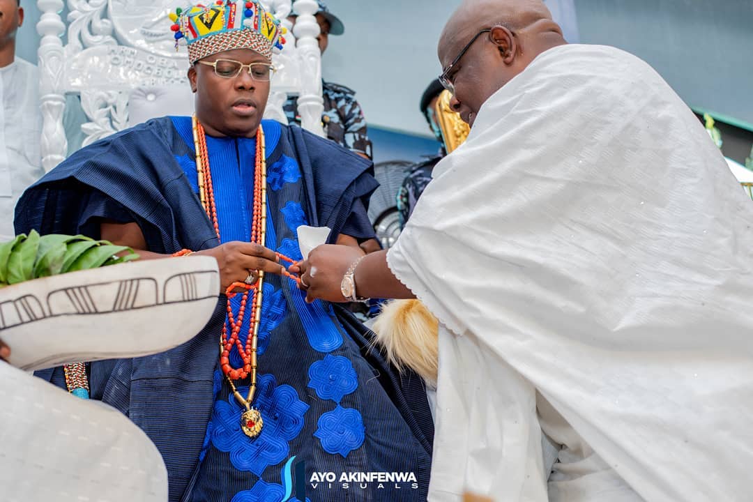 Man dressed in royal blue traditional attire and a colorful beaded crown receiving a ceremonial item from another person wearing white traditional clothing.