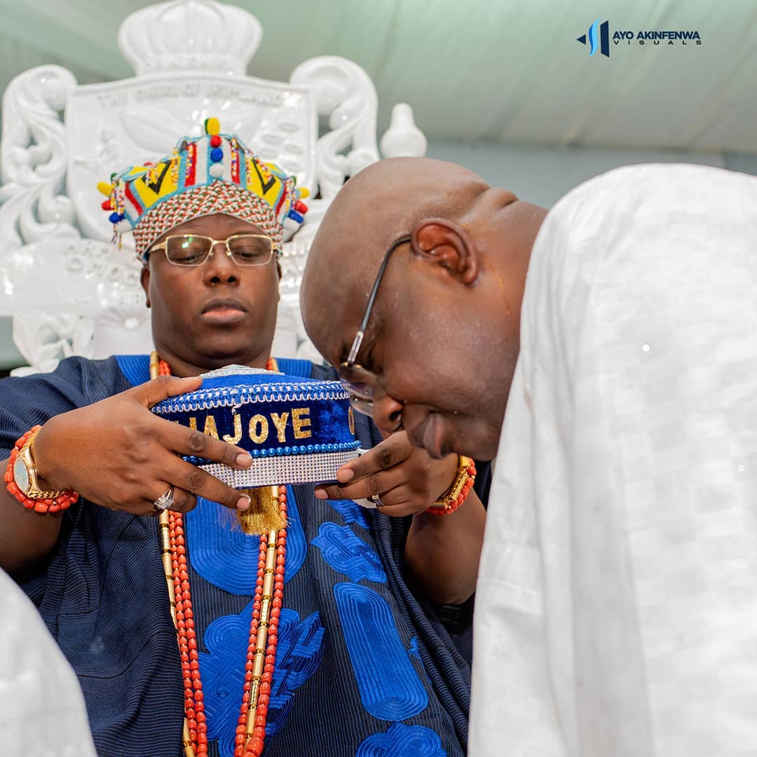 Man in traditional attire and colorful beaded crown holding a blue beaded crown inscribed with 'FIWAJOYE' while another man bows his head respectfully.