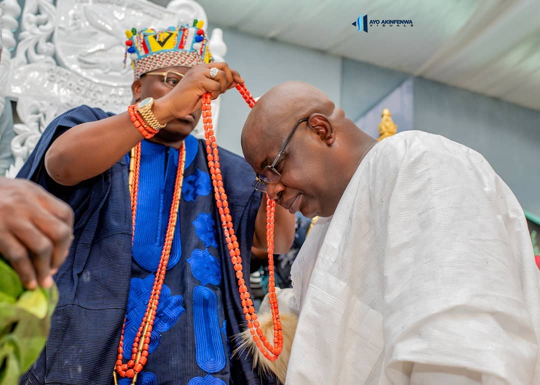 Man in traditional blue attire and colorful beaded crown placing a red coral bead necklace on a bowing man dressed in white traditional clothing and glasses.
