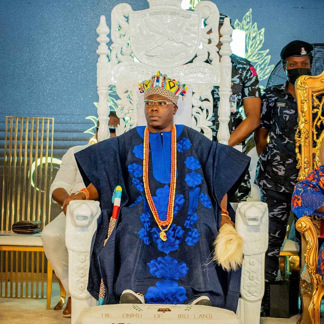 Man wearing traditional blue attire and beaded necklace sitting on ornate white throne with lion armrests.