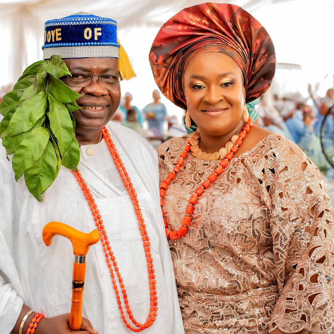 Smiling man and woman dressed in traditional Nigerian attire with coral bead necklaces, the man holds a wooden cane and wears a blue cap with leafy branches.