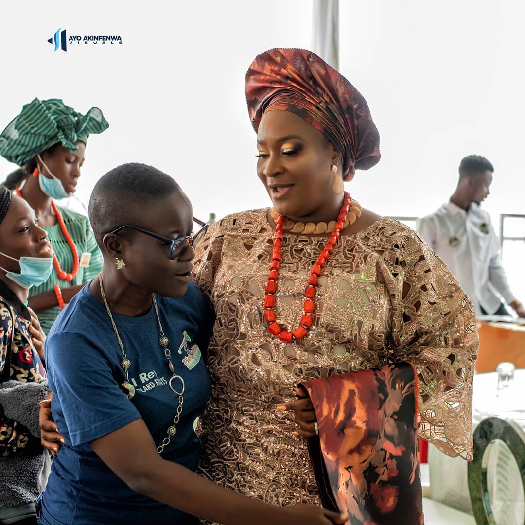Two women warmly embracing indoors, one wearing traditional attire with red beads and headwrap, the other in a blue t-shirt and glasses.