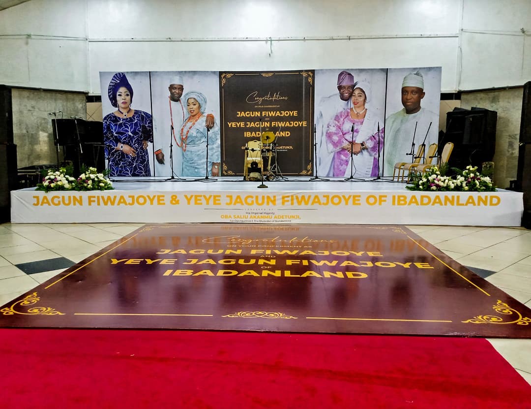 Indoor stage setup with large portraits of six people and text congratulating Jagun Fiwajoye and Yeye Jagun Fiwajoye of Ibadanland, decorated with floral arrangements and chairs.