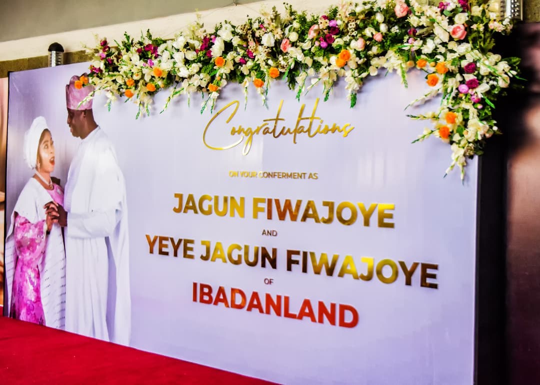 A man and woman in traditional attire standing beside a congratulatory banner decorated with flowers that reads congratulating Jagun Fiwajoye and Yeye Jagun Fiwajoye of Ibadanland.