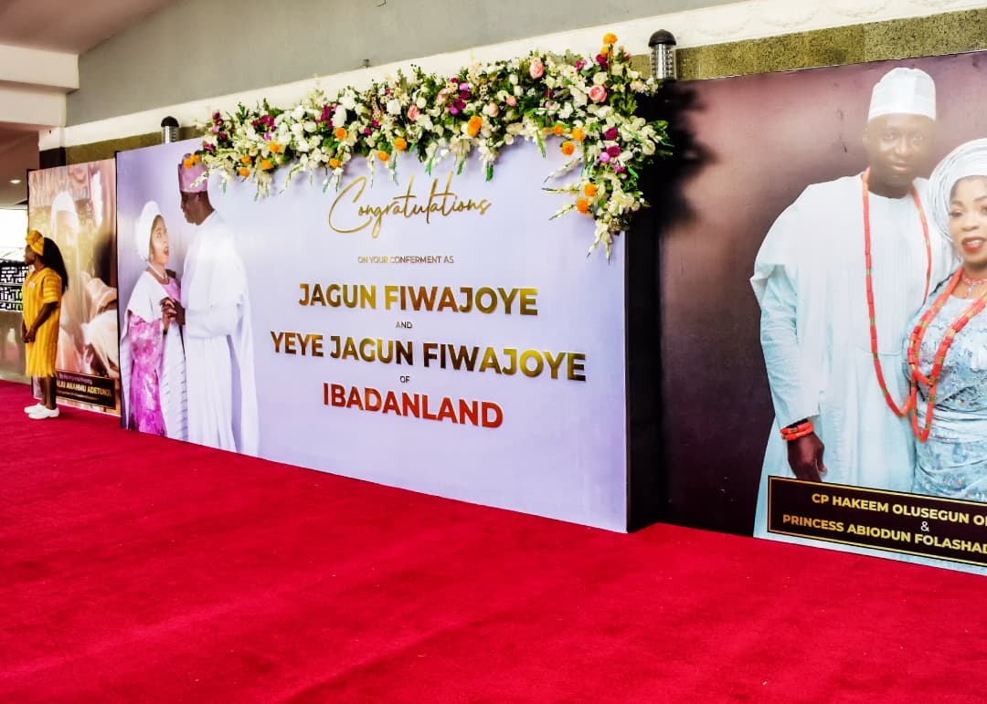 Decorated event backdrop with congratulatory message for Jagun Fiwajoye and Yeye Jagun Fiwajoye of Ibadanland, featuring floral arrangement and photos of couples.